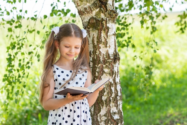 Una niña lee un libro en un día soleado de verano debajo de un árbol.