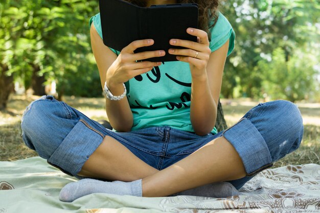 La niña lee el libro electrónico en un prado.