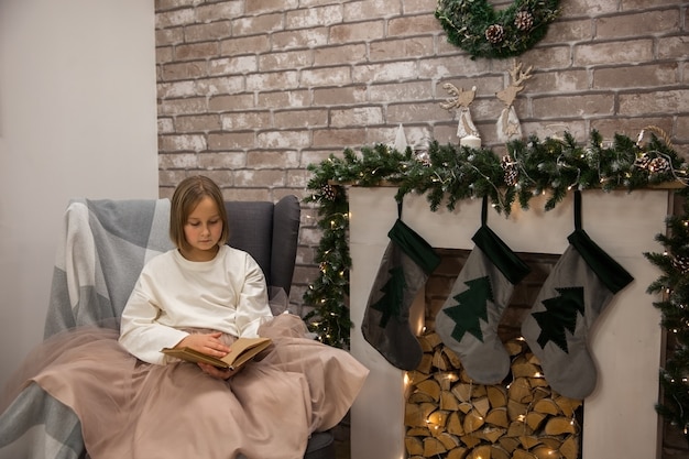 Una niña lee un libro en una silla junto a la chimenea, enfoque suave