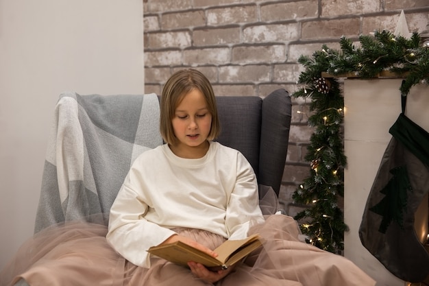 Una niña lee un libro en una silla junto a la chimenea, enfoque suave