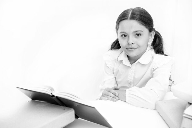 Niña leer libro sentarse mesa interior blanco Colegiala estudiando libro de texto Niño uniforme escolar cara feliz leer libro Emocionado por el conocimiento Concepto de educación en el hogar Libro interesante para niños