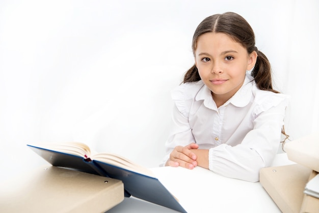 Niña leer libro sentarse mesa interior blanco. Colegiala estudiando libros de texto. Libro de lectura de cara feliz uniforme escolar para niños. Emocionado por el conocimiento. Concepto de educación en el hogar. Interesante libro para niños.