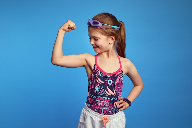 Niña levanta el brazo y mirando los músculos lleva gafas y traje de baño