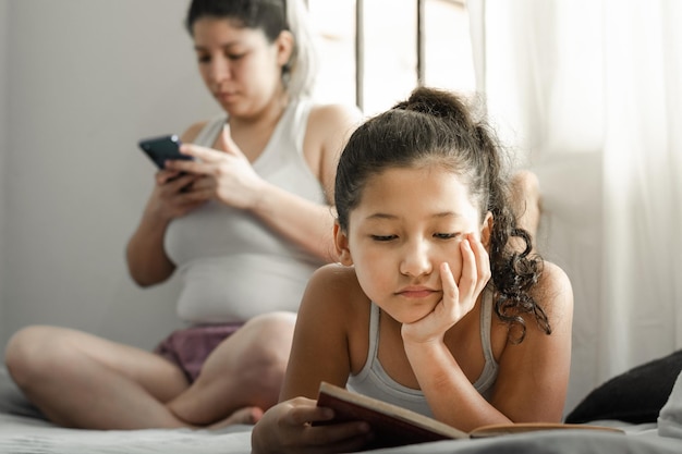 niña leyendo un libro acostado en su cama mientras su madre mira su teléfono celular