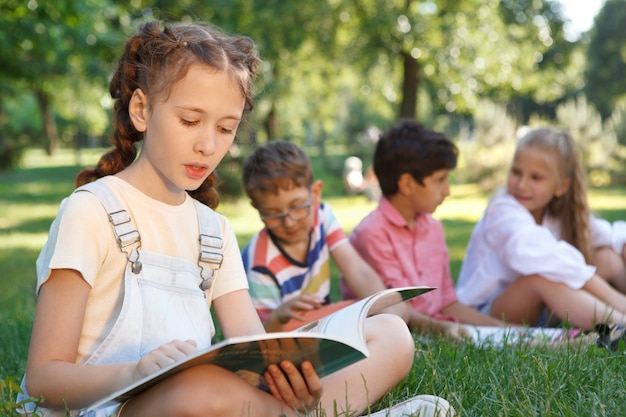 Niña leyendo un libro en el parque mientras sus amigos se relajan en el césped