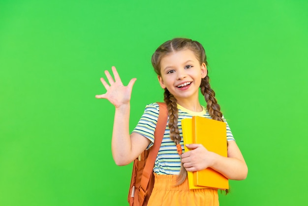 Una niña con libros en la mano y un maletín a la espalda agita la mano.