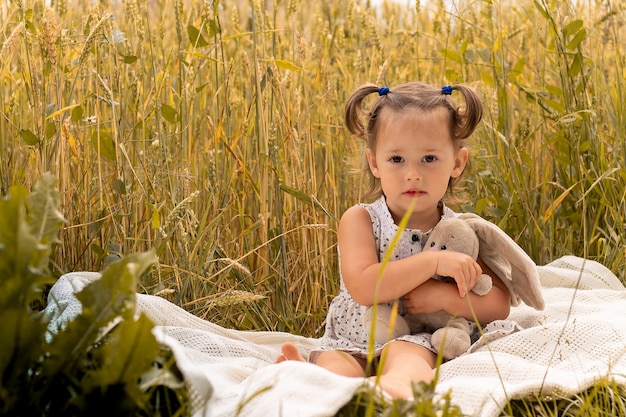 Niña linda 1-3 con un vestido ligero abraza a una liebre de peluche, sentada sobre un plaid en un campo de espiguillas de centeno en verano.