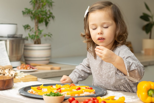 Niña linda 2-4 en vestido gris cocinar pizza en la cocina. Niño arregla los ingredientes en la base de la pizza