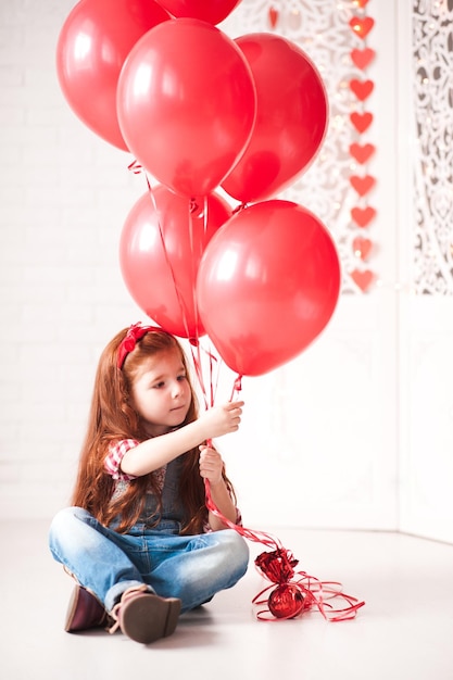 Niña linda de 4 a 5 años sosteniendo globos rojos sentados en la habitación Infancia infantil juguetona