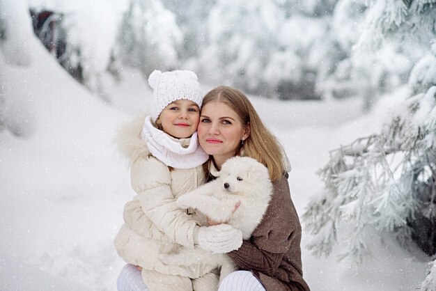 Niña linda de 4 años y su hermosa madre con un cachorro blanco esponjoso en el bosque de invierno