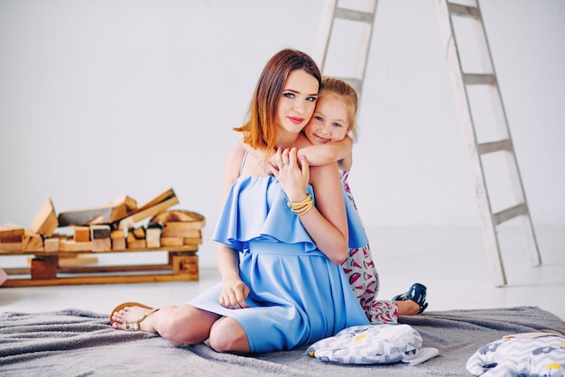 Niña linda abraza a su joven madre hermosa con amor. Hermosa madre con su hija en la habitación decorada en blanco.