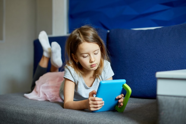 Niña linda acostada en el sofá, niño adicto a la tecnología, disfrutando de jugar juegos en línea en una tableta digital, usando aplicaciones