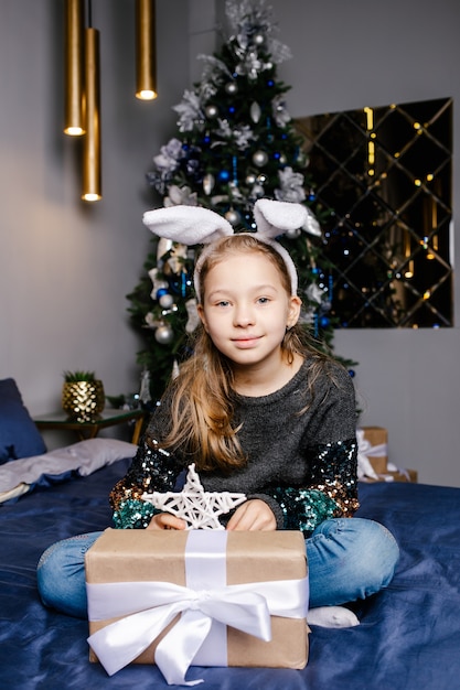 Niña linda alegre tiene una caja de regalo cerca del árbol de Navidad en el interior. Concepto de Navidad, vacaciones e infancia.