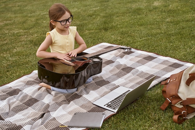 Niña linda con anteojos sentado sobre una hierba verde en el parque y sosteniendo el gasto de guitarra