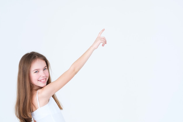 Niña linda apuntando a algo con la mano y el dedo índice en la pared blanca.