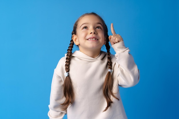 Niña linda apuntando hacia arriba y teniendo idea contra el fondo azul.