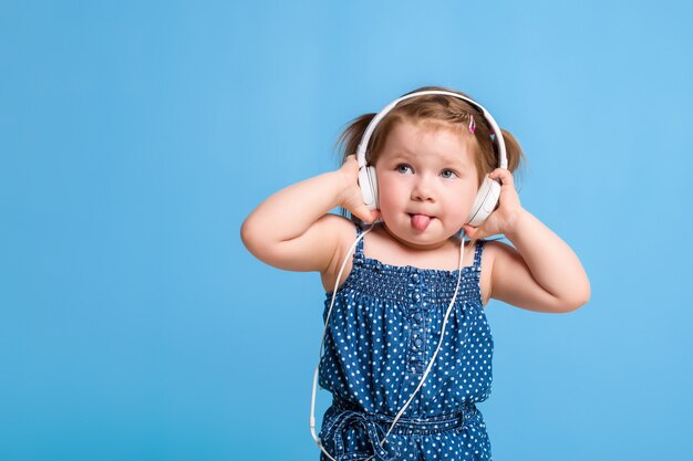 Foto niña linda en auriculares escuchando música con una tableta y sonriendo sobre fondo azul.