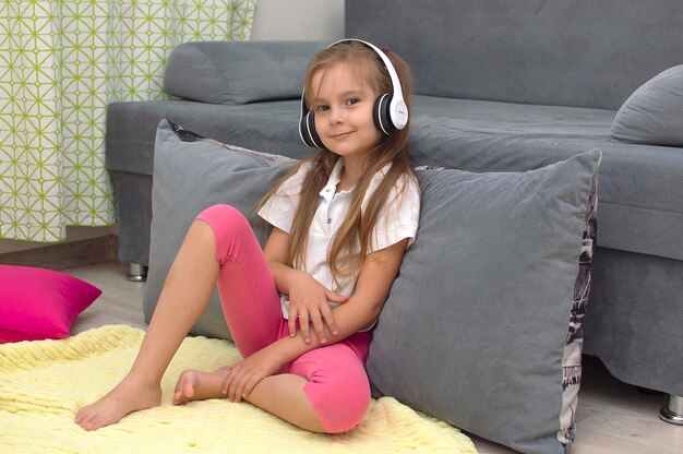 Una niña linda con auriculares está escuchando música, tirada en el suelo cerca del sofá gris de la sala de estar.