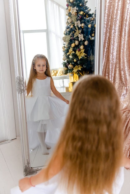 Niña linda con cabello largo hermoso se sienta cerca de un espejo y hace un peinado antes de Navidad.