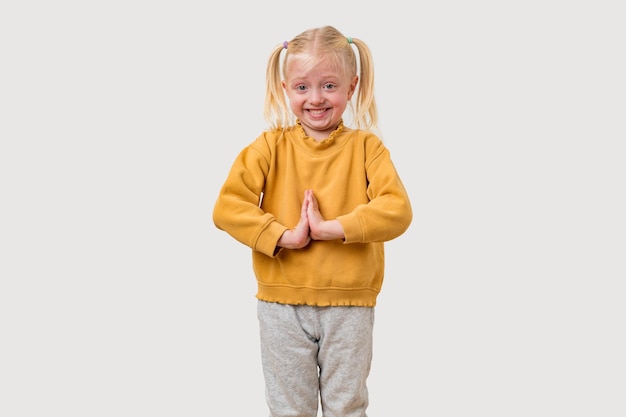 Una niña linda con cabello rubio, suéter amarillo y pantalones grises está haciendo yoga aislada sobre un fondo blanco.