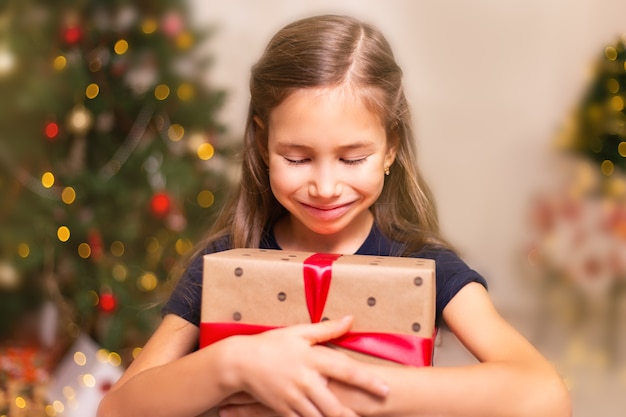 Niña linda con caja de regalo presente cerca del árbol de Navidad en casa.