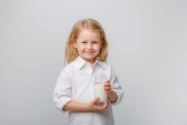 Niña linda con una camisa blanca sosteniendo un vaso de leche en un fondo blanco