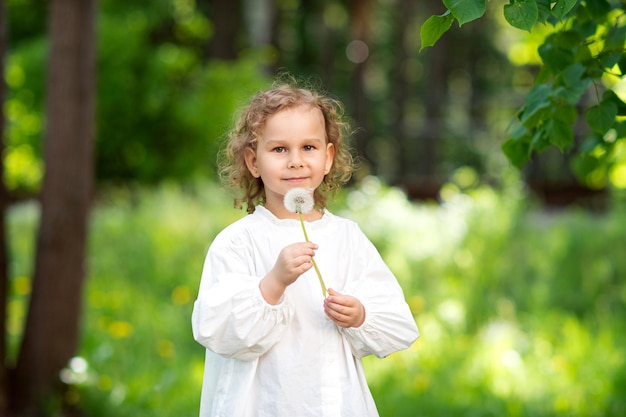 Una niña linda con una camisa blanca sostiene una flor de diente de león en sus manos y la sopla