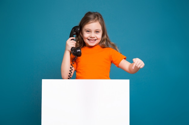 La niña linda en camiseta con el pelo marrón sostiene el papel limpio y habla por teléfono