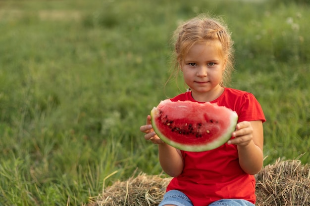 Niña linda con una camiseta roja y pantalones cortos de mezclilla con un trozo de sandía en las manos Un gran trozo de sandía roja madura en manos de una niña