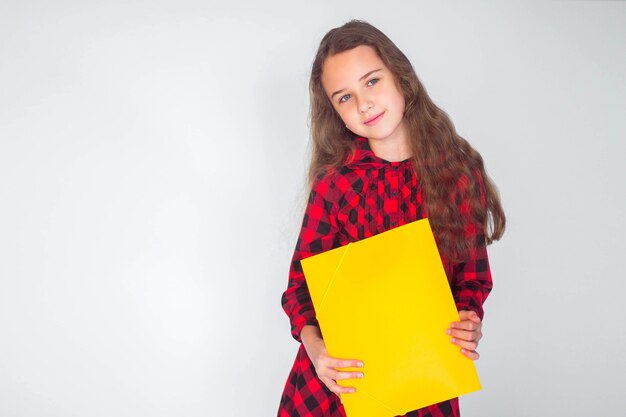 Niña linda con carpeta sonriendo de pie en la pared blanca, lugar para el texto, concepto de regreso a la escuela