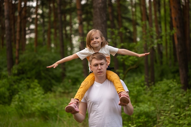 Niña linda caucásica sentada sobre los hombros de su padre en el bosque. Padre e hija jugando juntos, riendo y divirtiéndose. Concepto de actividad familiar feliz