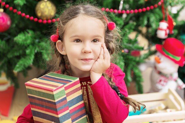 Niña linda cerca del hermoso árbol de Navidad con un regalo