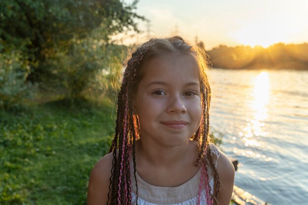 Niña linda con coletas africanas en el fondo del lago y la playa en primer plano mira misteriosamente a la cámara. Hermosa puesta de sol en el parque junto al río. Infancia feliz. Vacaciones escolares.