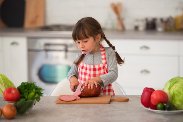 Niña linda con coletas en un delantal culinario a cuadros corta salchichas en sándwiches en la cocina en la mesa.