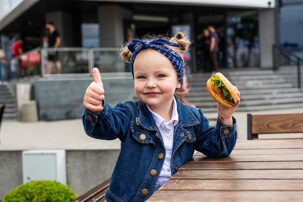 Niña linda comiendo una hamburguesa mostrando los pulgares para arriba en un café. Concepto de comida rápida para niños