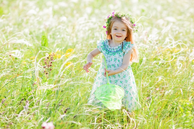 Niña linda con una corona de flores en la cabeza camina en un campo y atrapa red de mariposas