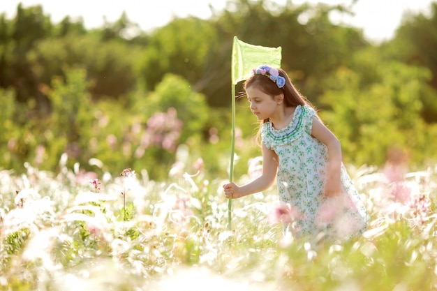 Niña linda con una corona de flores en la cabeza camina en un campo y atrapa red de mariposas