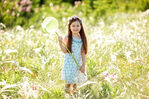 Niña linda con una corona de flores en la cabeza camina en un campo y atrapa red de mariposas