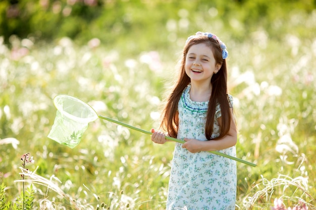 Niña linda con una corona de flores en la cabeza camina en un campo y atrapa red de mariposas