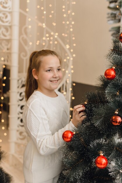 Niña linda decora el árbol de Navidad con bolas de juguetes rojos. Una chica con un vestido de suéter de punto blanco coloca bolas colgantes en un abeto artificial