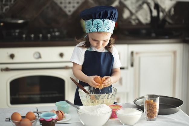 Niña linda en un delantal y un gorro de cocinero rompe un huevo en un plato