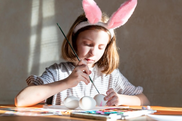 Niña linda con diadema de orejas de conejo pintando huevos de Pascua en la mesa en el interior Bricolaje para Pascua con niños Artesanía para vacaciones de primavera Decoraciones para colorear huevos para vacaciones Concepto de creatividad