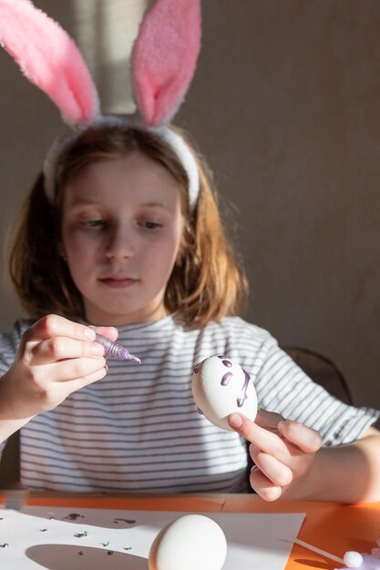 Niña linda con diadema de orejas de conejo pintando huevos de Pascua en la mesa en el interior Bricolaje para Pascua con niños Artesanía para vacaciones de primavera Decoraciones para colorear huevos para vacaciones Concepto de creatividad
