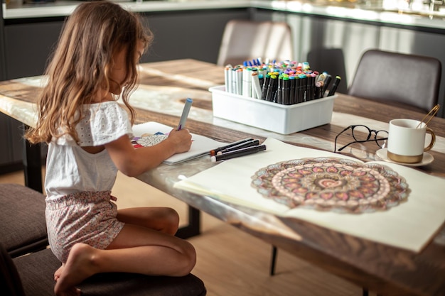 Una niña linda dibuja un patrón de mandala circular en el álbum con marcadores de arte.