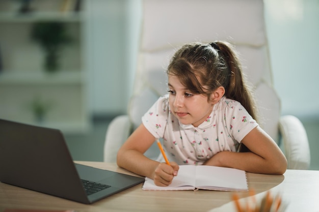 Una niña linda y diligente que usa su computadora portátil para hacer una lección en línea en casa.