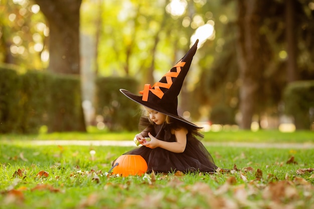 Una niña linda con un disfraz de bruja camina por la calle con un caramelo en forma de calabaza.