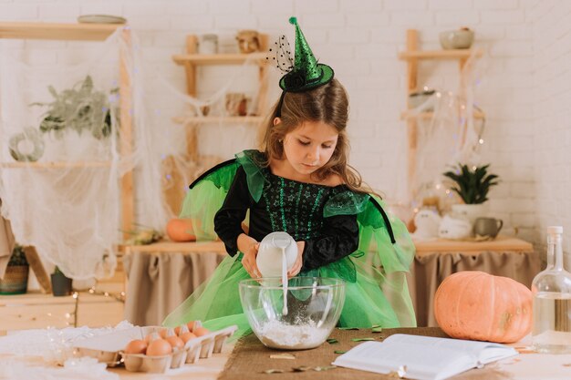 Niña linda en un disfraz de Halloween verde de una bruja o un hada prepara un pastel de calabaza