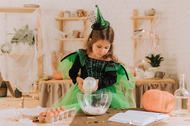 Niña linda en un disfraz de Halloween verde de una bruja o un hada prepara un pastel de calabaza