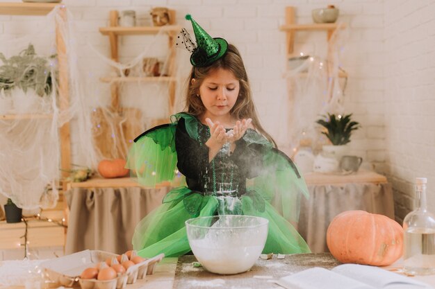 Niña linda en un disfraz de Halloween verde de una bruja o un hada prepara un pastel de calabaza