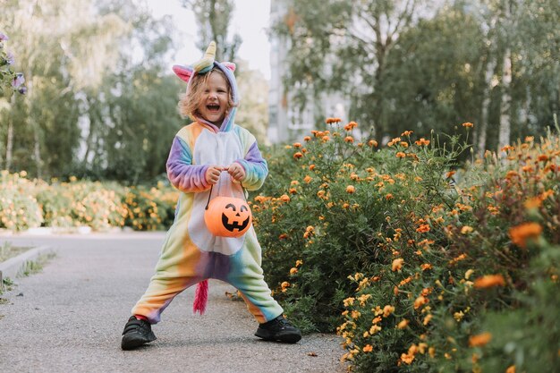 Niña linda con un disfraz de unicornio arcoíris para Halloween va a recolectar dulces en una canasta de calabazas